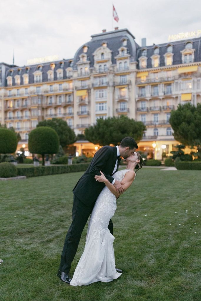 Man and woman kiss on the grounds of a grand hotel in Switzerland, as part of their wedding day.