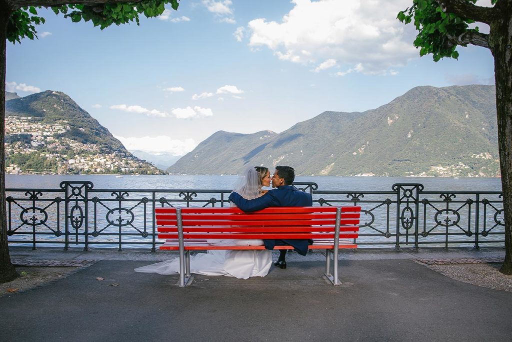 Newlywed couple sits on a red bench overlooking the lake on the grounds of the Hotel Splendide Royal, Lugano, Switzerland.