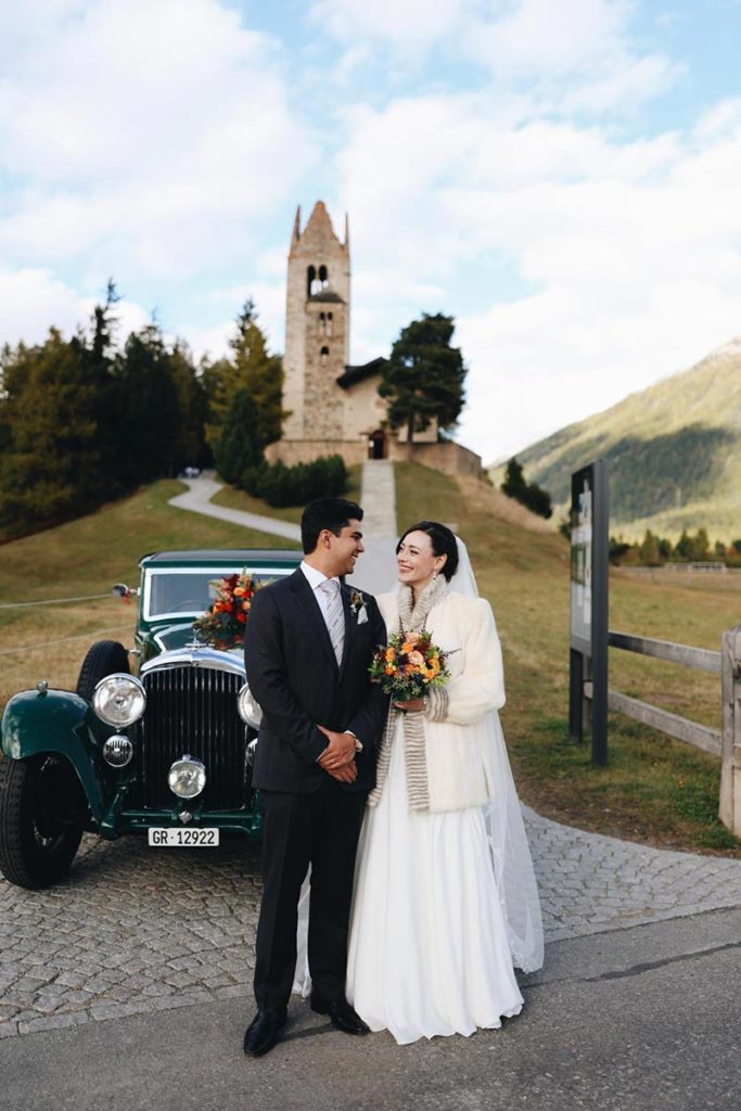 Bride in her wedding dress and a coat holding her bridal bouquet poses with her husband in front of the historic San Gian Church in St. Moritz.