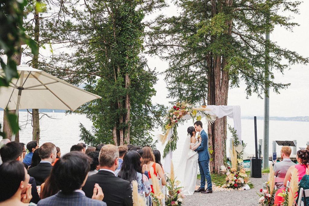 Bride and groom kiss during their outdoor wedding ceremony at the Hotel Vitznauerhof with Lake Lucerne in the background.