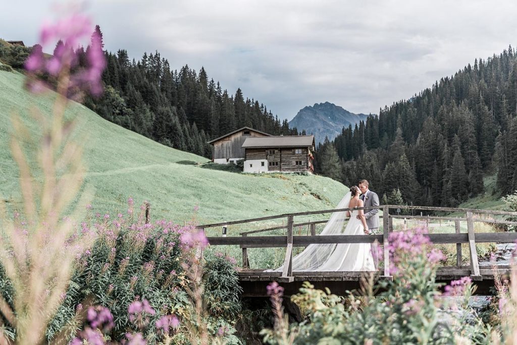 Bride and groom standing on a bridge kissing in the Sertig Valley with an older wooden building in the background.