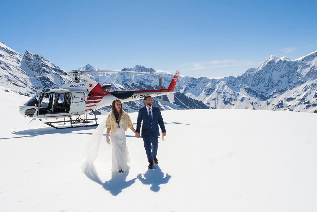 Wedding couple walks on a Swiss glacier with a helicopter in the background, as part of their destination Swiss wedding.